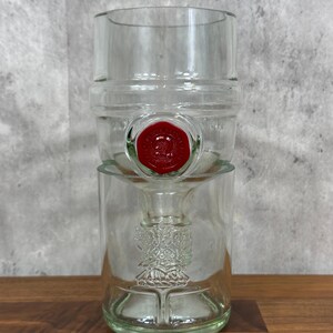 May include: Clear glass bottle with a red seal. The bottle has a unique design with a wide top and a narrower base. The glass has a slight green tint. The bottle is sitting on a wooden surface.
