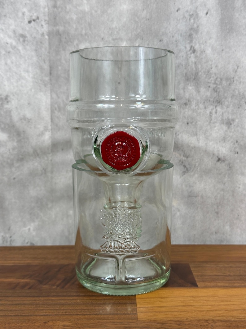 May include: Clear glass bottle with a red seal. The bottle has a unique design with a wide top and a narrower base. The glass has a slight green tint. The bottle is sitting on a wooden surface.