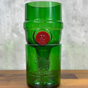 May include: Two-piece green glass set, likely repurposed from a bottle. The top piece is barrel-shaped with a red seal. The bottom piece is a cylindrical glass with an embossed design. The set is displayed on a wooden surface.