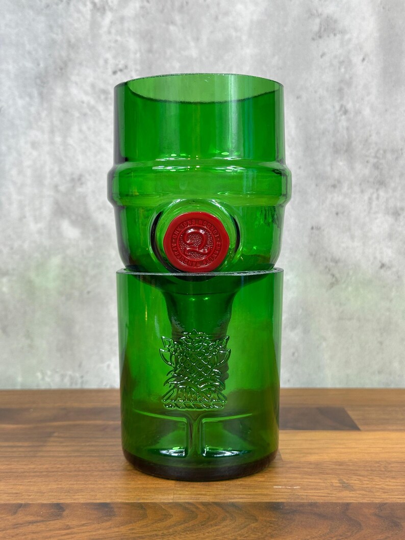 May include: Two-piece green glass set, likely repurposed from a bottle. The top piece is barrel-shaped with a red seal. The bottom piece is a cylindrical glass with an embossed design. The set is displayed on a wooden surface.