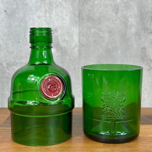 May include: A green glass bottle and tumbler set. The bottle has a red and white seal and a short neck. The tumbler features a tree design. Both items are made of recycled glass and sit on a wooden surface.