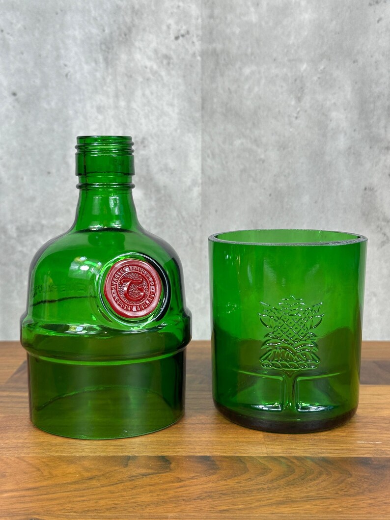 May include: A green glass bottle and tumbler set. The bottle has a red and white seal and a short neck. The tumbler features a tree design. Both items are made of recycled glass and sit on a wooden surface.