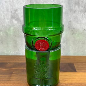 May include: Two green glass tumblers stacked on top of each other. The top tumbler has a red seal. The bottom tumbler has a decorative design. The tumblers are made from recycled glass.