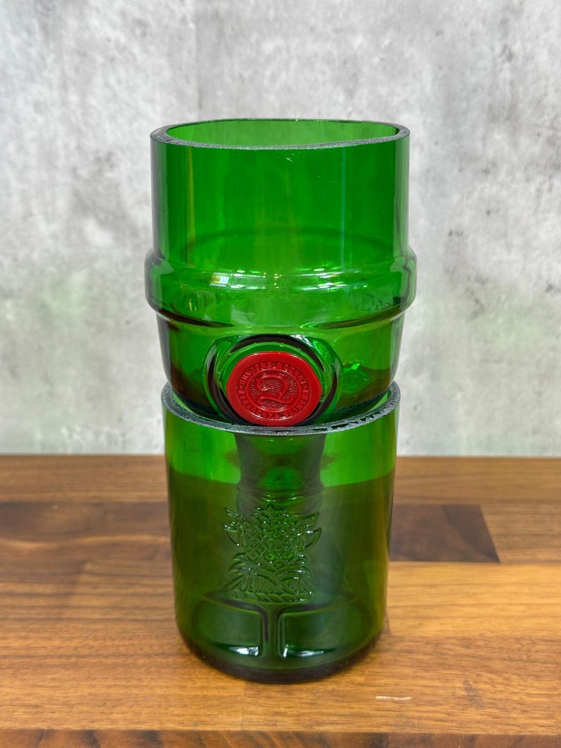 May include: Two green glass tumblers stacked on top of each other. The top tumbler has a red seal. The bottom tumbler has a decorative design. The tumblers are made from recycled glass.