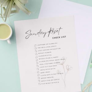 May include: A white checklist titled "Sunday Reset" with a handwritten font. The checklist includes items like "Gratitude or Journaling" and "Tidy Main Living Spaces." A cup of green liquid and a plant are in the background.