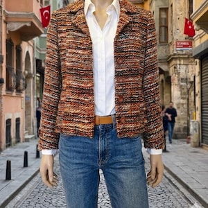 Puede incluir: Una blazer de tweed multicolor con cinturón de cuero marrón y vaqueros azules. La blazer tiene cierre de botones y cuello. El atuendo se usa sobre una camisa blanca abotonada. El fondo muestra una calle adoquinada.