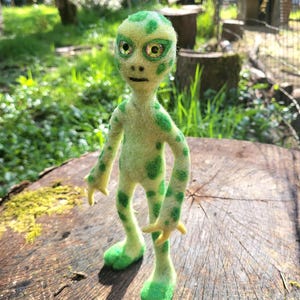 May include: A handmade felt alien figure in shades of green. The alien has large, expressive eyes, a small mouth, and green spots on its body. It stands on a wooden surface, with a natural outdoor background.