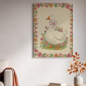 Puede incluir: Impresión en lienzo enmarcada que presenta un ganso blanco con pico y patas naranjas, adornado con un lazo rosa. El ganso se encuentra entre flores, enmarcado por un borde floral con flores rosas y naranjas. El fondo tiene una textura.