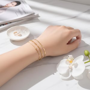 Peut inclure: Un ensemble de trois bracelets dorés portés au poignet. Le lot comprend une fine chaîne avec une petite perle, une chaîne à maillons texturés d'épaisseur moyenne et une chaîne serpent plate et lisse. Les bracelets sont polis et brillants.