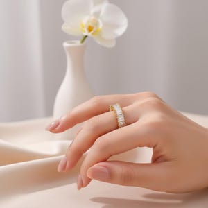 Peut inclure: Une bague dorée ornée d'une rangée continue de pierres transparentes rectangulaires taillées en baguette, fixées par des griffes. La bague est portée au doigt, mettant en valeur la disposition uniforme des pierres scintillantes sur l'anneau.
