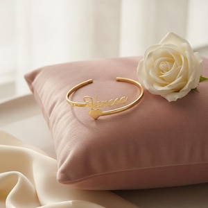 Peut inclure: Un bracelet manchette en métal doré arborant le prénom Vanesa en écriture cursive et un petit pendentif en forme de cœur à l'extrémité ouverte. Le bracelet présente une finition polie et un design circulaire simple ouvert sur le devant.