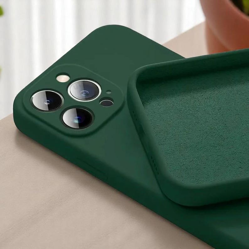 May include: A dark green phone case with a matte finish. The case is shown with the camera lens cutouts visible. The interior of the case is a lighter shade of green. The case is designed to protect the phone.