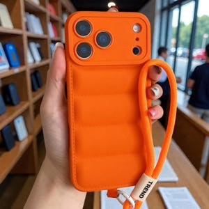 Peut inclure: Coque de téléphone orange vif avec un motif matelassé et une lanière assortie. L'étui comporte un module de caméra surélevé avec trois objectifs et un flash. Le mot "TREND" est imprimé sur la lanière.