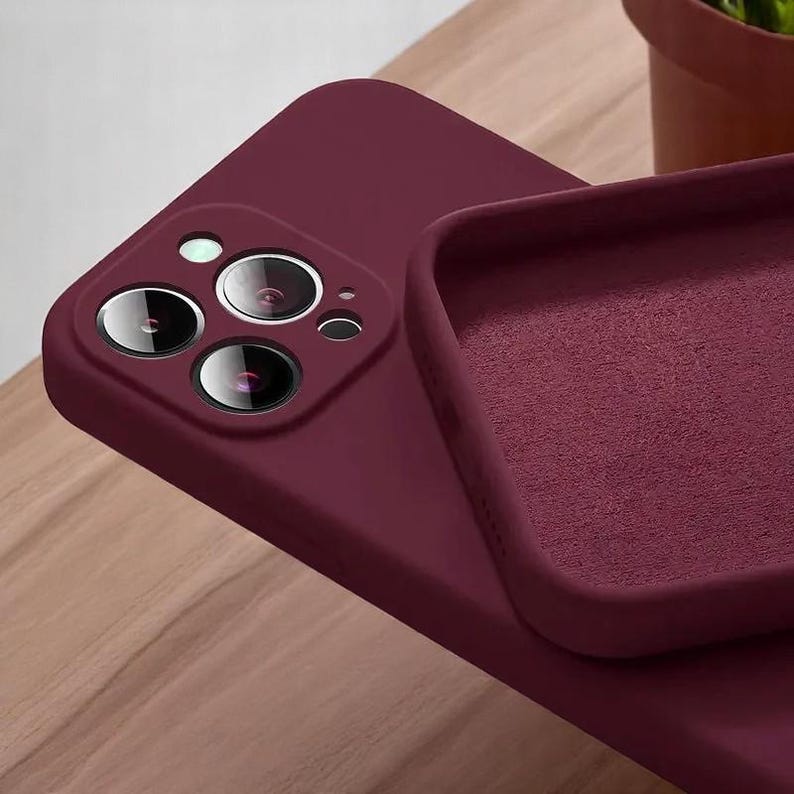 May include: A burgundy-colored phone case with a soft, matte finish. The case is shown with the phone, highlighting the camera lenses. The interior of the case is also burgundy. The phone case is designed to protect the phone.