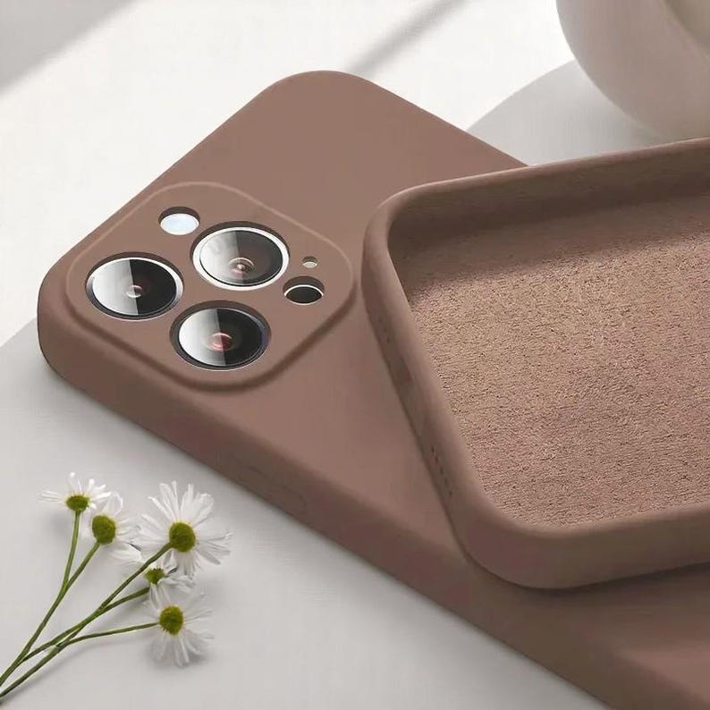 May include: A close-up of a brown phone case with a camera lens cutout. The case is made of a soft material and has a raised edge. White daisy flowers are in the lower left corner.