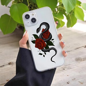 May include: Clear phone case featuring a black snake coiled around two red roses with green leaves. The design is printed on a clear case, allowing the phone's color to show through. The case has a glossy finish.