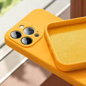 May include: Close-up of a bright yellow phone case. The case has a soft, matte finish and features cutouts for the phone's camera lenses. The interior of the case is a matching yellow color. The case is designed to protect the phone.
