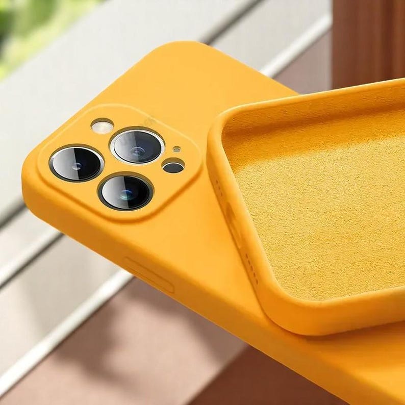 May include: Close-up of a bright yellow phone case. The case has a soft, matte finish and features cutouts for the phone's camera lenses. The interior of the case is a matching yellow color. The case is designed to protect the phone.