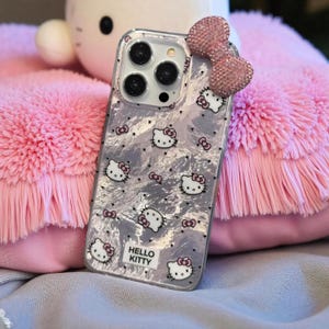 K&ouml;nnte beinhalten: Transparente Handyh&uuml;lle mit silbernem Marmormuster und Hello Kitty-Grafiken. Die H&uuml;lle hat eine rosa Schleife und ein Hello Kitty-Logo. Die H&uuml;lle liegt auf einem flauschigen rosa Kissen.