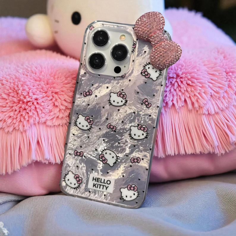 K&ouml;nnte beinhalten: Transparente Handyh&uuml;lle mit silbernem Marmormuster und Hello Kitty-Grafiken. Die H&uuml;lle hat eine rosa Schleife und ein Hello Kitty-Logo. Die H&uuml;lle liegt auf einem flauschigen rosa Kissen.