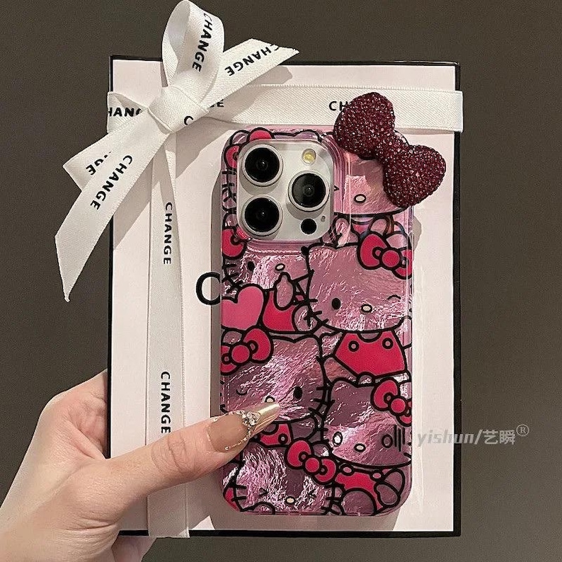 K&ouml;nnte beinhalten: Rosa Handyh&uuml;lle mit Hello Kitty-Design, Schleifen und Herzen. Die H&uuml;lle hat einen transparenten Rand und eine dekorative Schleife. Pr&auml;sentiert mit einem wei&szlig;en Band und einer Geschenkbox.