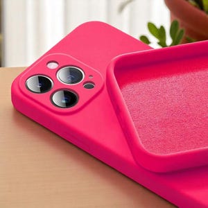 May include: A close-up of a bright pink phone case, showcasing the camera lenses and the interior lining. The case is made of a smooth material and appears to be designed for a smartphone. The case is on a light brown surface.