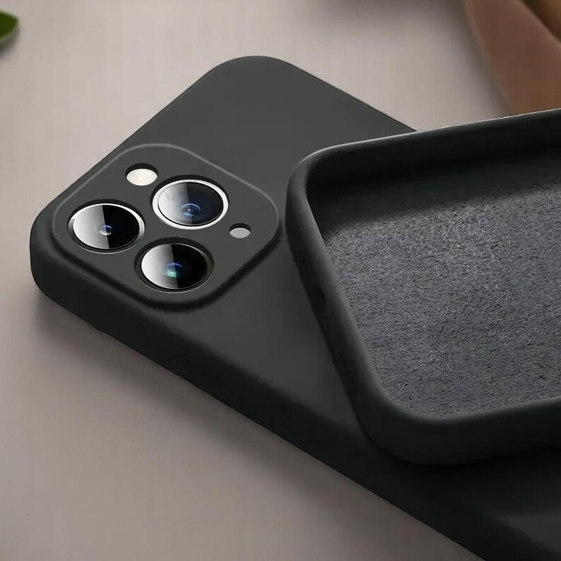 May include: A black phone case with a matte finish, featuring a triple-lens camera cutout. The case is shown partially open, revealing a soft, gray interior lining. The phone case is designed to protect the phone.