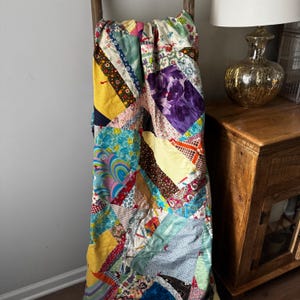 May include: A colorful patchwork quilt draped over a wooden ladder. The quilt features a variety of fabric patterns and colors, including yellow, blue, purple, and floral prints. The quilt is a mix of textures and designs.