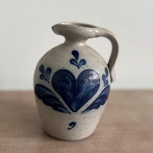 May include: A small, stoneware pitcher with a handle and spout. The pitcher is beige with a blue heart and leaf design. The heart is in the center of the pitcher, with leaves on either side. The handle is curved and attached to the top of the pitcher.