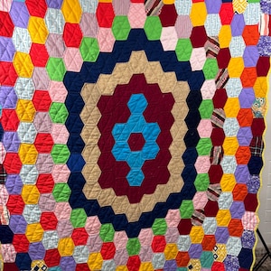 May include: A large, rectangular quilt constructed from numerous small, multicolored fabric hexagons stitched together in a honeycomb pattern. The design features a central bright blue cross shape surrounded by concentric rings of maroon, tan, and navy.