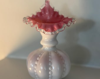May include: A white and pink glass vase with a ruffled rim. The vase has a bulbous body with vertical beaded accents. The rim is a vibrant pink with a clear, ruffled edge. The vase is likely vintage or antique.