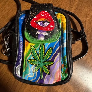 Puede incluir: Un pequeño bolso bandolera de cuero negro con un diseño psicodélico colorido. El bolso presenta un hongo pintado con un ojo, una hoja de marihuana y un fondo de arcoíris.