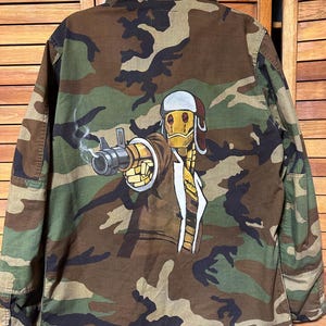 Könnte beinhalten: Tarnfarbene Militärjacke mit einer handgemalten Illustration auf dem Rücken. Das Kunstwerk zeigt einen Robotercharakter, der eine Waffe hält, mit Details in Gelb, Braun und Weiß. Die Jacke hat ein klassisches Tarnmuster in Grün-, Braun- und Schwarztönen.