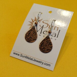 May include: A pair of copper dangle earrings with a black swirl design. The earrings are on a white card with the text "Sun Metal Jewelry" and the website address "www.SunMetalJewelry.com".