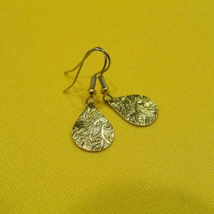 May include: A pair of gold-tone earrings with a teardrop shape and a textured, swirling pattern. The earrings are on a yellow background.