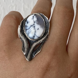 Może przedstawiać: Srebrny pierścionek z owalnym kamieniem z czarno-białym wzorem dendrytycznym. Kamień osadzony jest w teksturowanej srebrnej oprawie o organicznym kształcie. Pierścionek jest na dłoni.