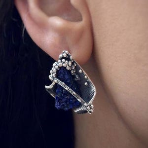 May include: A silver earring featuring a dark blue stone and small white details. The earring, with an abstract leaf design, is worn on an ear. The design combines various textures and shapes, creating a unique accessory. The earring is a statement piece.