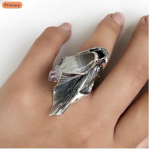 May include: A silver-toned statement ring featuring a large, irregularly shaped stone with gray and white patterns. The ring has a detailed, leaf-like design on the band and a smaller purple stone accent. The ring is worn on a finger.