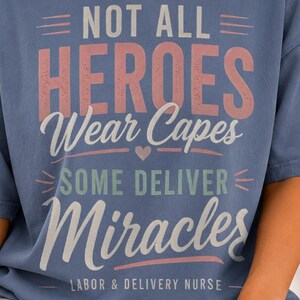 Könnte beinhalten: Ein blaues Kurzarm-T-Shirt mit dem Text "NOT ALL HEROES Wear Capes SOME DELIVER Miracles LABOR & DELIVERY NURSE" in rosa, weißer und hellblauer Schrift sowie einem kleinen Herzsymbol, das mittig zwischen den Textzeilen platziert ist.