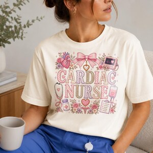 Könnte beinhalten: Ein cremefarbenes T-Shirt mit einem Grafikdruck, der den Text CARDIAC NURSE in einem Karomuster zeigt. Das Design enthält Illustrationen eines menschlichen Herzens, einer rosa Schleife, eines Herzmonitors, eines Stethoskops und Blumen.