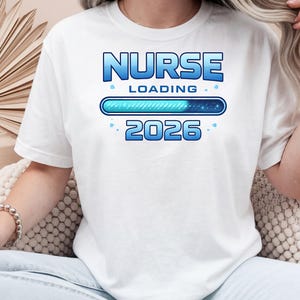Könnte beinhalten: Weißes T-Shirt mit einem blauen Aufdruck "NURSE LOADING 2026". Das Design beinhaltet einen Ladebalken und Sterne. Das T-Shirt besteht aus einem weichen Material.