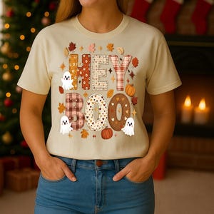 Könnte beinhalten: Beiges T-Shirt mit dem Schriftzug "HEY BOO" in einem Patchwork-Design, mit Herbstblättern, Kürbissen und Geisterillustrationen. Das Shirt wird mit einer blauen Jeans kombiniert.