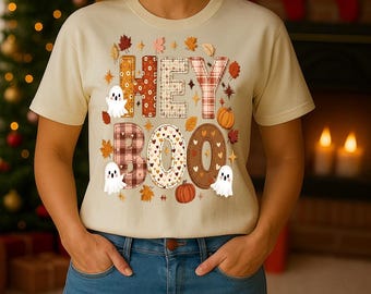 May include: Beige t-shirt with the words "HEY BOO" in a patchwork design, featuring fall leaves, pumpkins, and ghost illustrations. The shirt is paired with blue jeans.