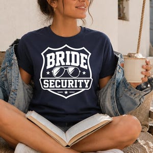 Könnte beinhalten: Marineblaues T-Shirt mit weißem Aufdruck „BRIDE SECURITY“ in einem Schild-Design, mit Sonnenbrille und Sternen. Die Person trägt eine Jeansjacke und hält eine Tasse und ein Buch.