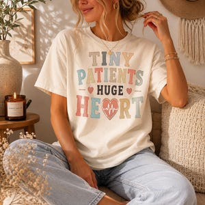Könnte beinhalten: Ein cremefarbenes Kurzarm-T-Shirt mit dem Text "TINY PATIENTS HUGE HEART" in mehrfarbigen Blockbuchstaben. Das Design enthält kleine rote Herzen und ein zentrales Herzmotiv mit einem weißen EKG-Herzschlagmuster in der Mitte.