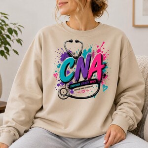 Könnte beinhalten: Ein beiges Langarm-Sweatshirt mit einem Grafikdruck, der den Text CNA, Compassion, Strength, Impact und Certified Nursing Assistant zeigt. Das Design enthält ein schwarzes Stethoskop, bunte Farbspritzer und kleine Herzsymbole.
