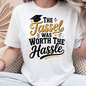Könnte beinhalten: Weißes T-Shirt mit dem Text "The Tassel Was Worth The Hassle" in Schwarz und Gold. Das Design beinhaltet eine Abschlusskappe und Sterne, ideal für eine Abschlussfeier.