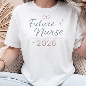 Könnte beinhalten: Weißes T-Shirt mit dem Text "Future Nurse 2026" in Schreibschrift. Das Design beinhaltet eine Krankenschwestermütze und kleine Sterne. Das Shirt hat einen Rundhalsausschnitt und scheint aus weichem Material zu sein.