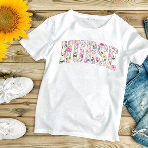 May include: White t-shirt with a floral print design that spells out the word "NURSE". The shirt is laid out on a wooden surface with sunflowers, white sneakers, and blue jeans.