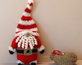Pode incluir: Uma figura de Pai Natal em croché com um gorro pontiagudo às riscas vermelhas e brancas, barba branca encaracolada, fato vermelho com cinto preto de fivela dourada e botas pretas. A figura segura um saco dourado texturado por um cordão.